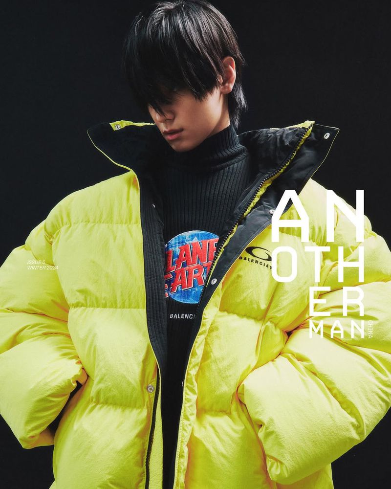 Adult male fashion portrait in a puffy yellow coat and black turtleneck sweater with a graphic print against a dark studio backdrop.