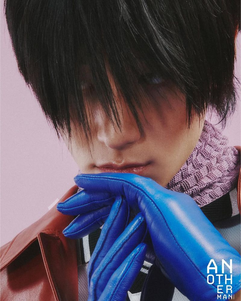 Close-up portrait of a person with black hair and blue gloves, wearing a red jacket and a patterned scarf against a pink background.