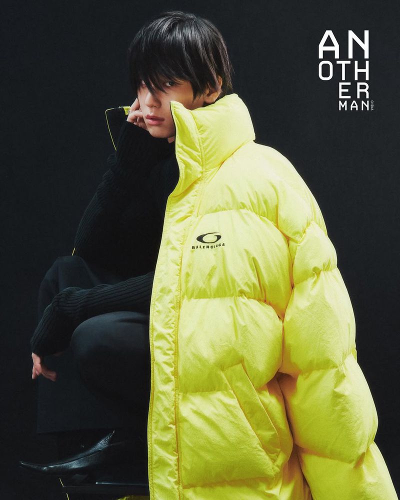 Portrait of a young adult with dark hair wearing a yellow puffer jacket and dark clothing against a dark grey studio backdrop.