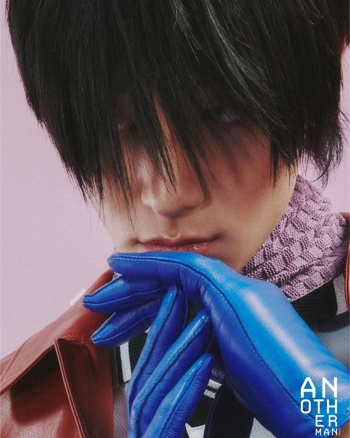 Close-up portrait of a person wearing blue leather gloves, a textured purple scarf, and a red jacket against a pink backdrop