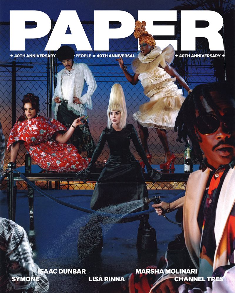 Paper magazine cover features a diverse group of models in high fashion against a chain-link fence backdrop with water spraying on the ground.