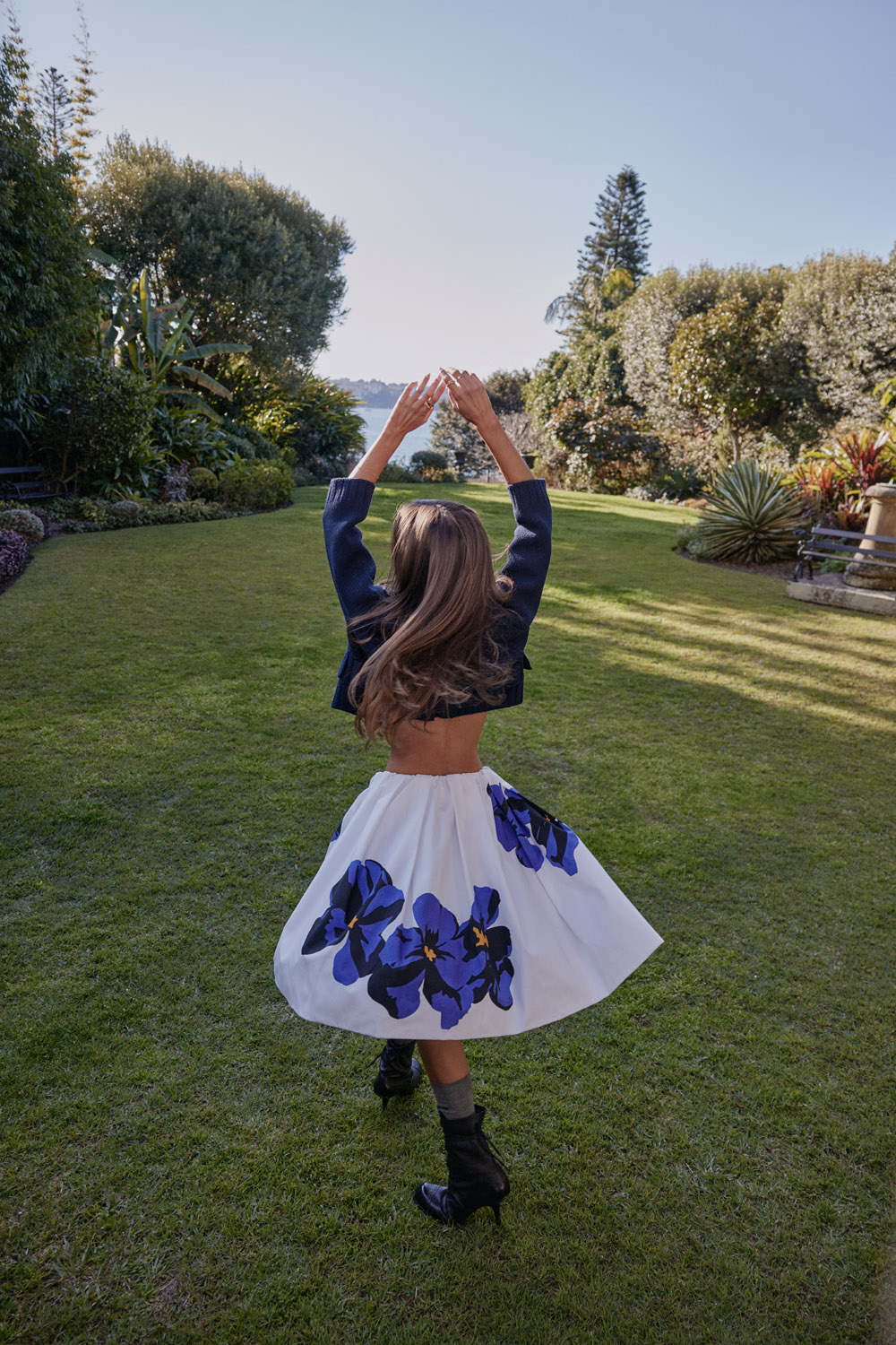 Young woman in floral skirt twirls with arms raised in lush garden, back to camera, black jacket and boots, sunny day.