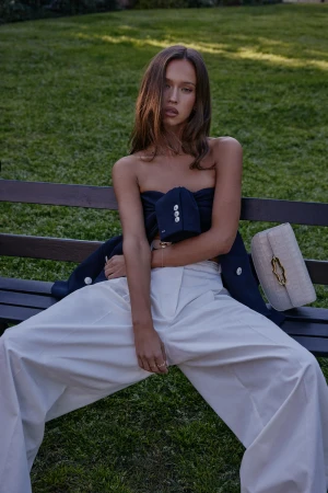 Brunette woman in stylish blue and white fashion sitting on a park bench with greenery, natural light, and a fashion purse.
