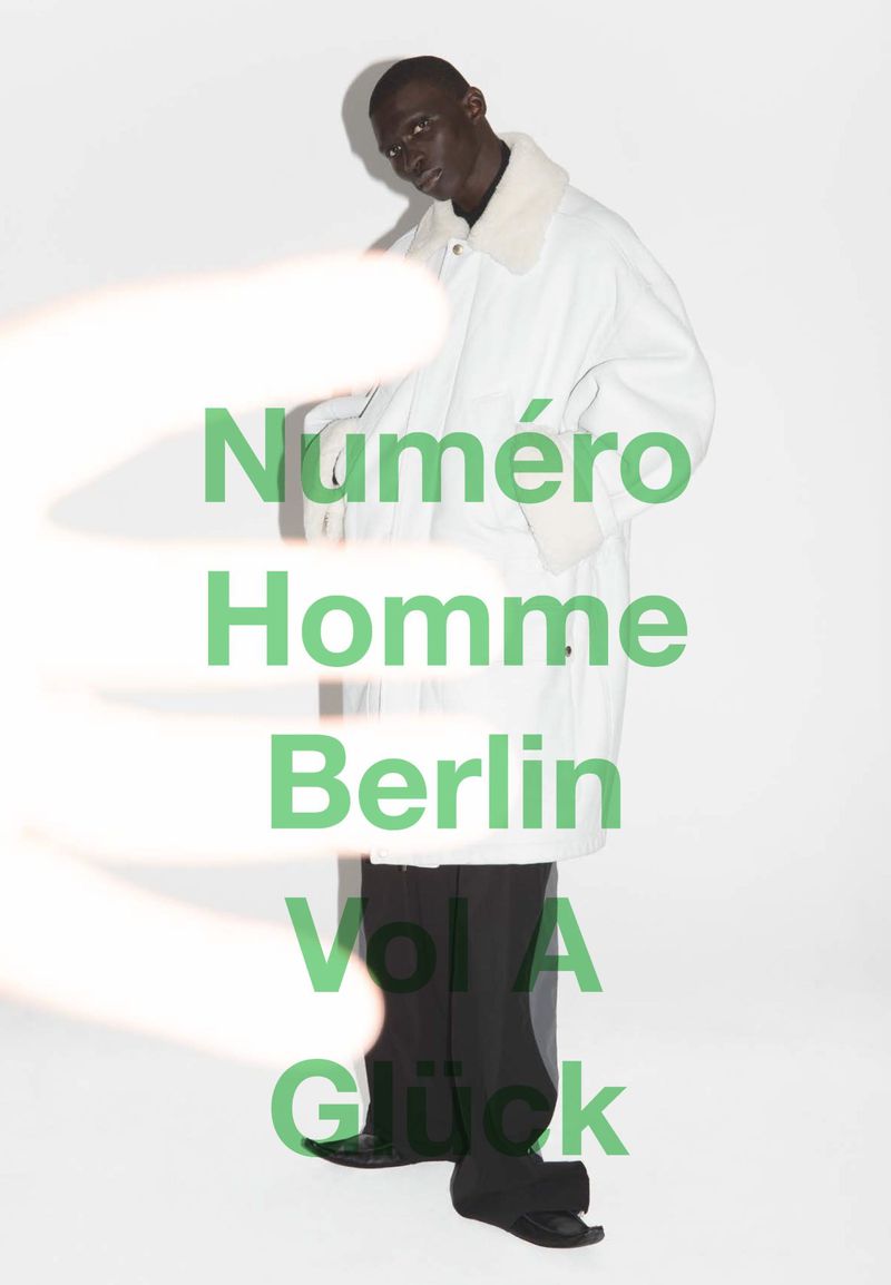 Editorial fashion shot of a Black male model wearing a white coat and black pants in a photo studio, with a hand obstructing the view.