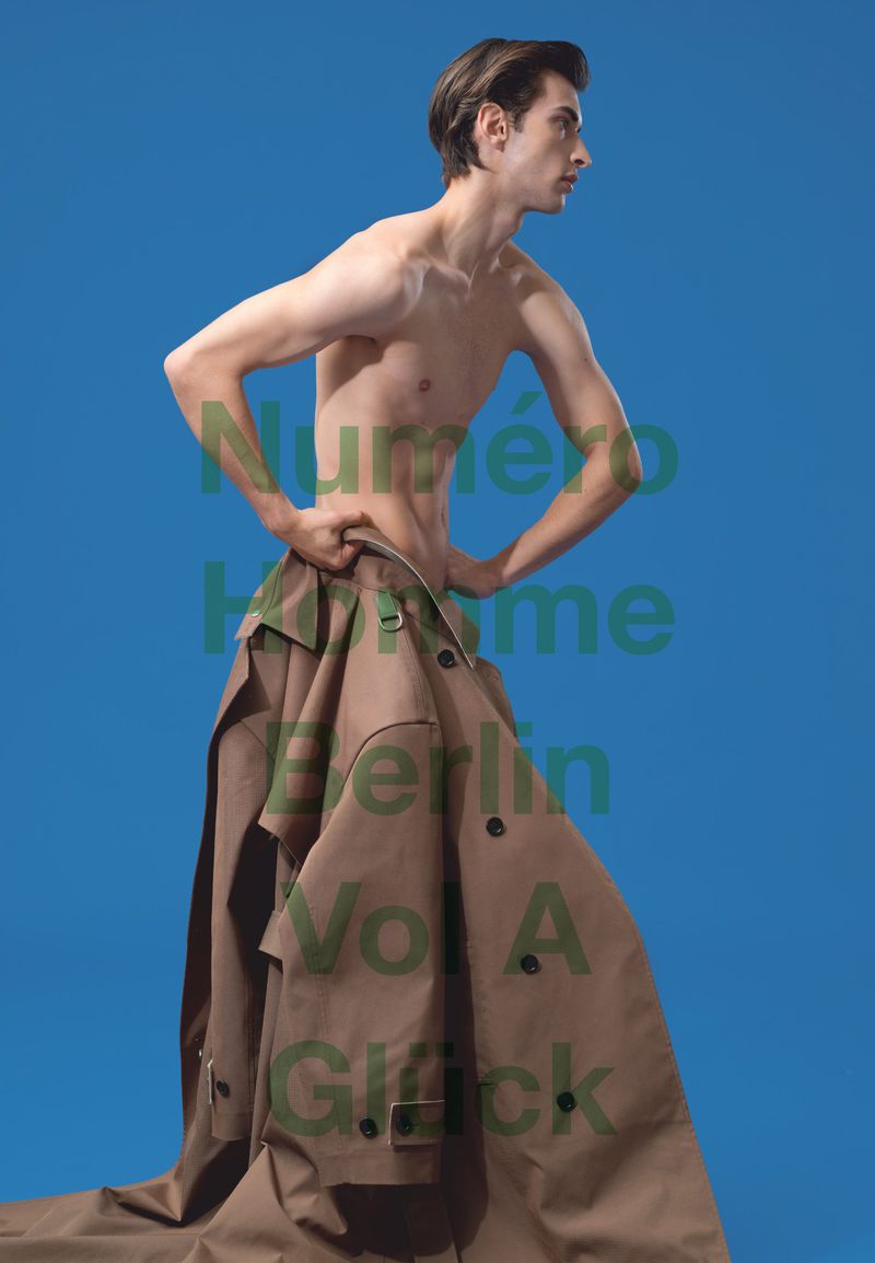 Male fashion model poses in front of a blue background. He is wearing a long brown trench coat slung around his lower half.