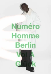 Black man wearing a white coat with a fur collar standing against a plain white background with foreground hand blur.
