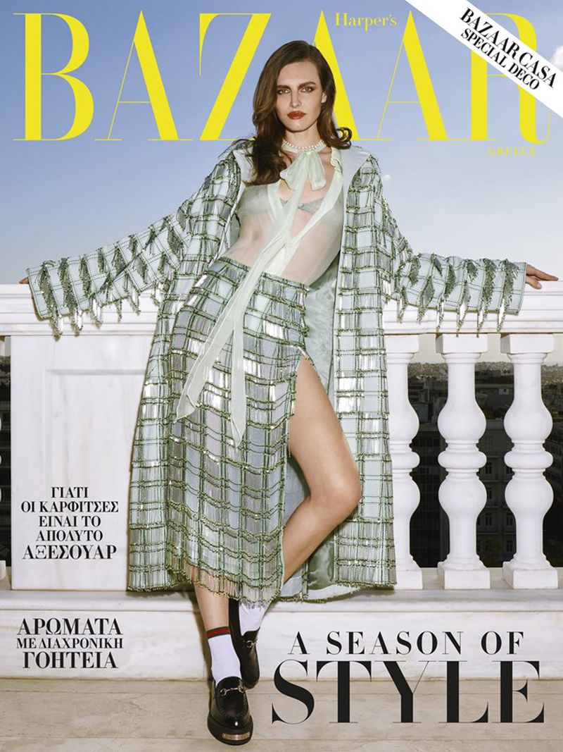 Bazaar magazine cover featuring a model in a plaid skirt and coat posing on a balcony against a blue sky background.