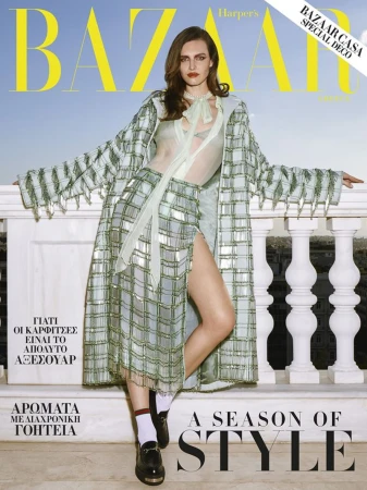 Model leaning against a white balustrade wearing a patterned green coat, matching skirt, white socks, and loafers against an open sky.