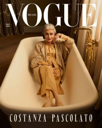 Elderly woman with grey hair seated inside a white vintage bathtub wearing gold garments for Vogue Brasil December 2024