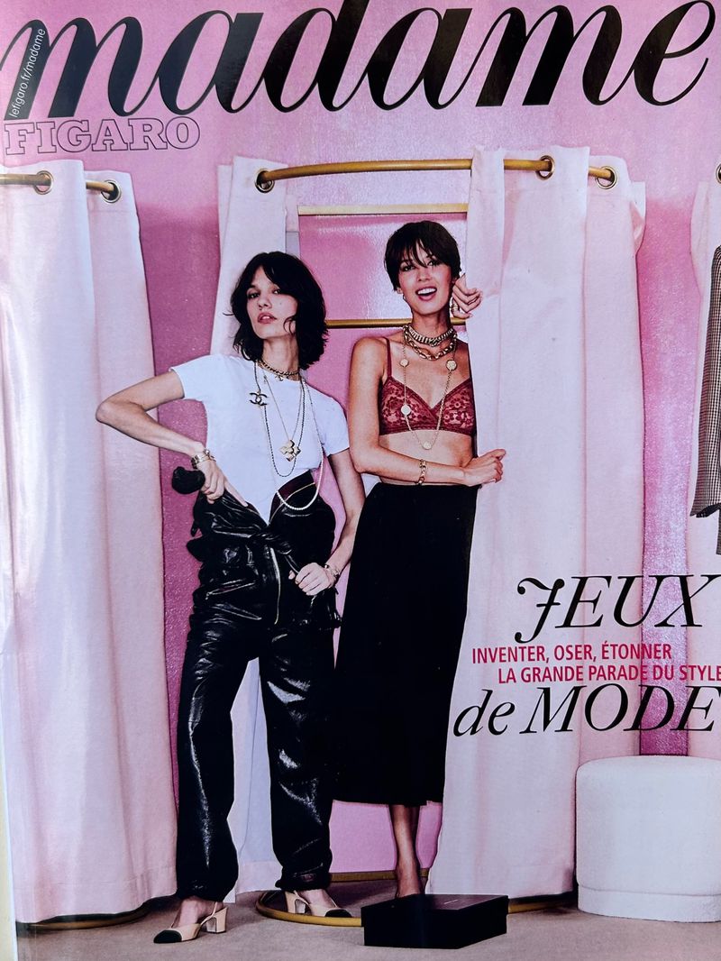 Two fashion models pose in front of a pink dressing room setup for a magazine cover, showcasing stylish clothing and accessories.