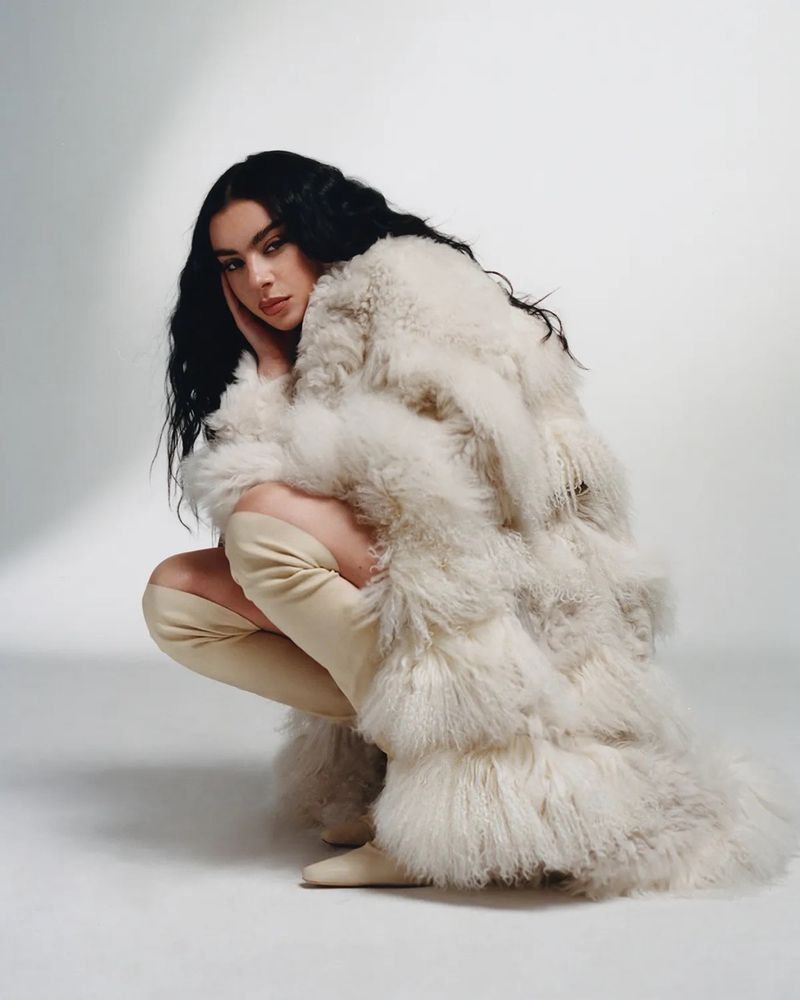 A woman with dark hair poses in a squatting position wearing a cream colored, textured coat and matching thigh high boots in a studio.