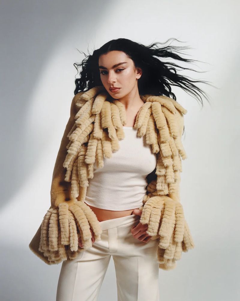 Brunette woman in fringed coat poses in a studio. She wears a white crop top and white pants with a neutral studio background.