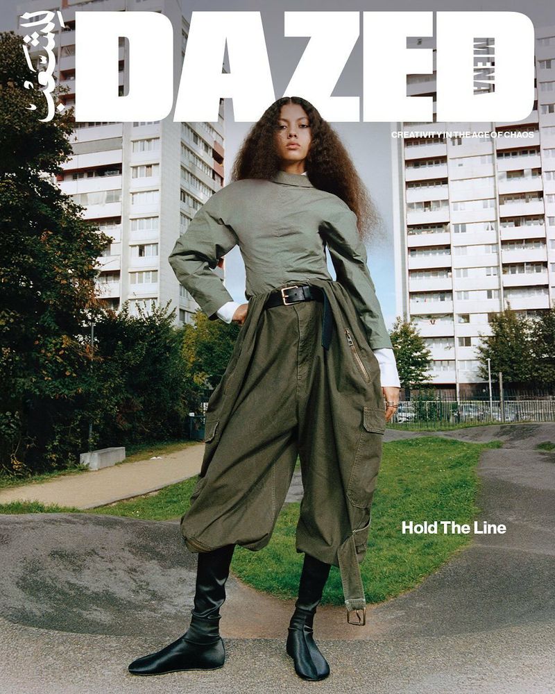 Editorial fashion shot of a young adult woman with curly hair wearing an olive green top, baggy pants, and black boots in an urban setting.