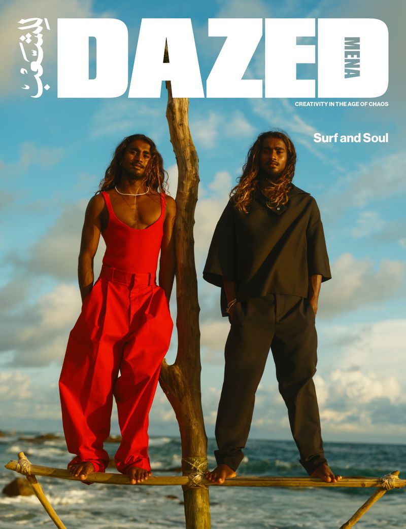 Two men pose on a wooden structure by the ocean, one in red, one in black, under a blue sky, for a magazine cover.