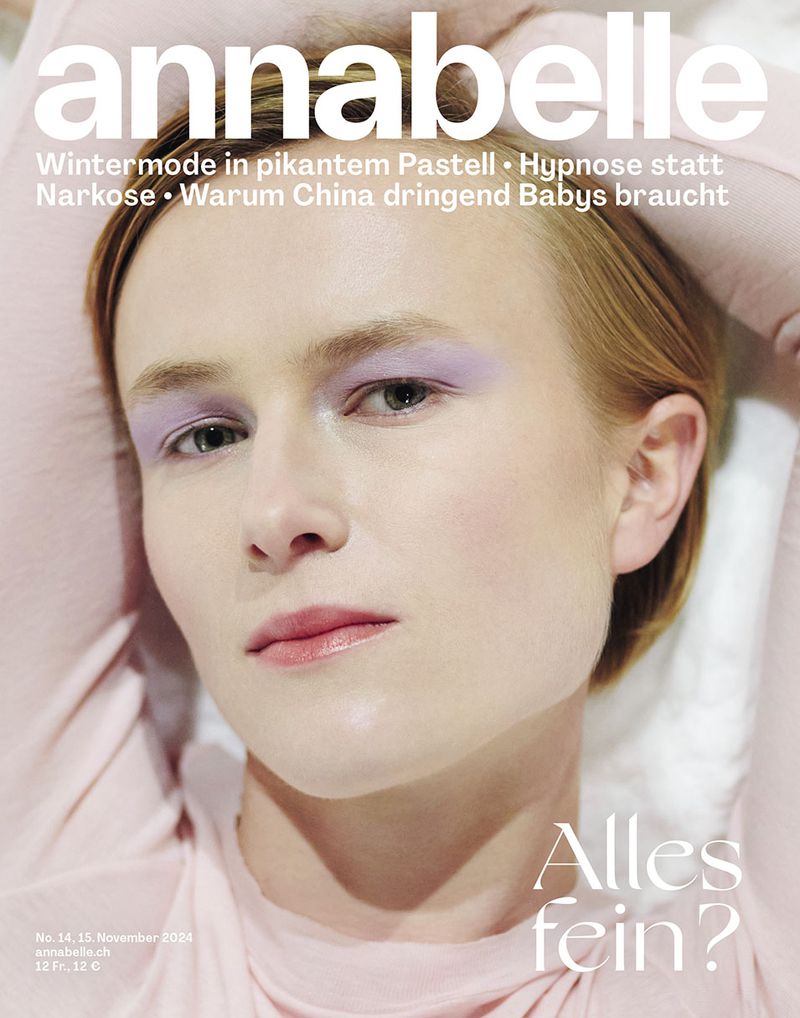 Close-up magazine cover featuring a model with light skin, blonde hair, and light purple eyeshadow, wearing a pink top. Simple studio lighting.