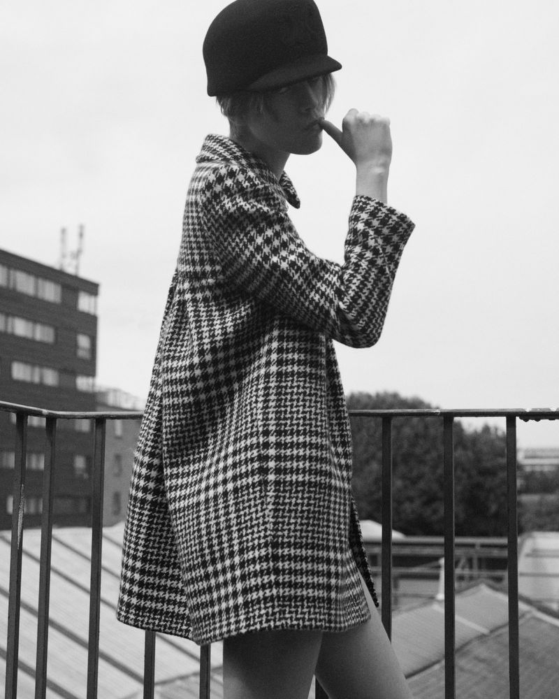 Model wearing a houndstooth coat and dark cap standing on a rooftop with a metal railing against an overcast sky