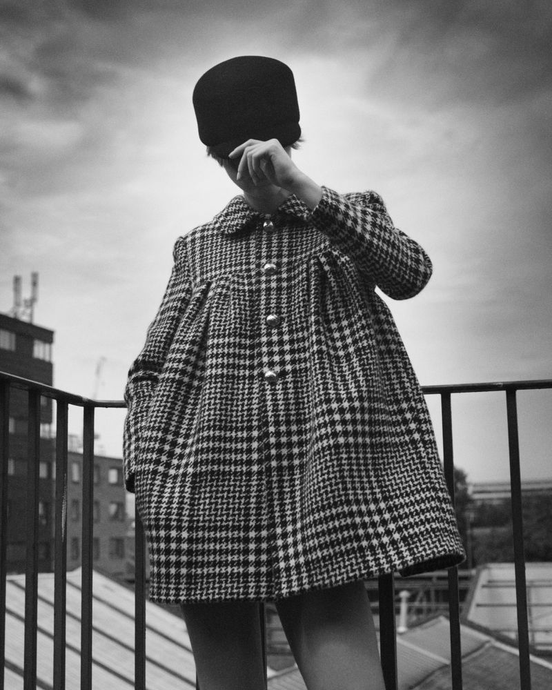 Model wearing a houndstooth coat and dark cap standing against a cloudy sky on a rooftop for Crosscurrent #5.