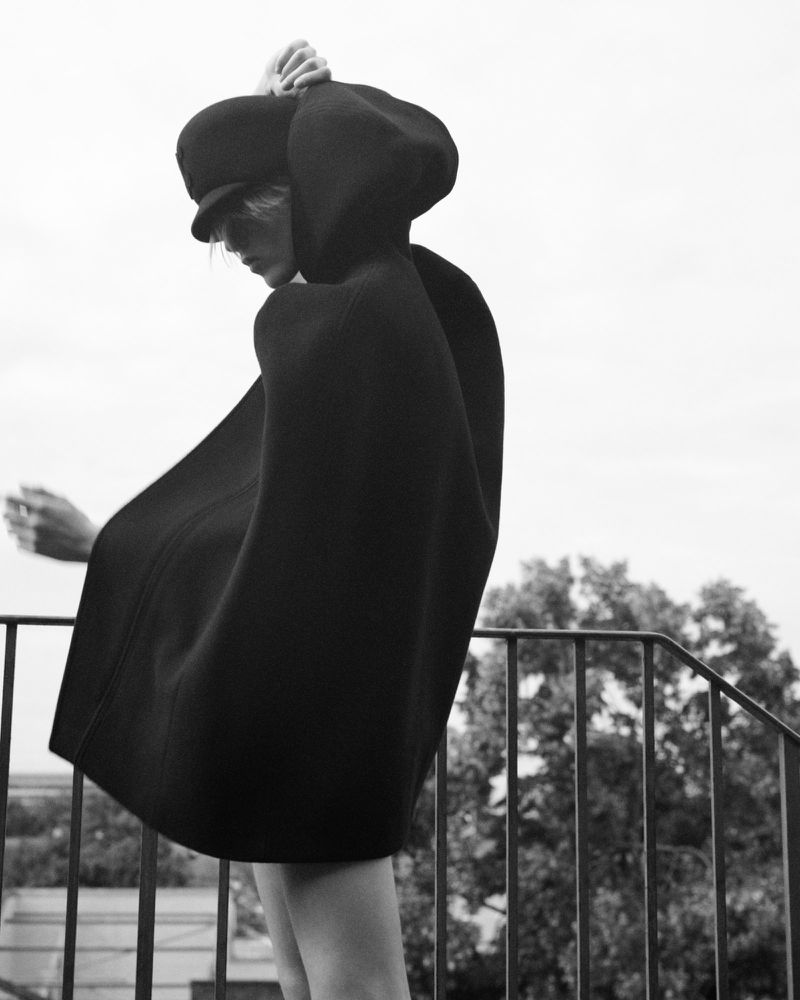 Monochrome portrait of a person wearing a hooded cape and cap on a balcony, with trees visible in the background, against a grey sky.