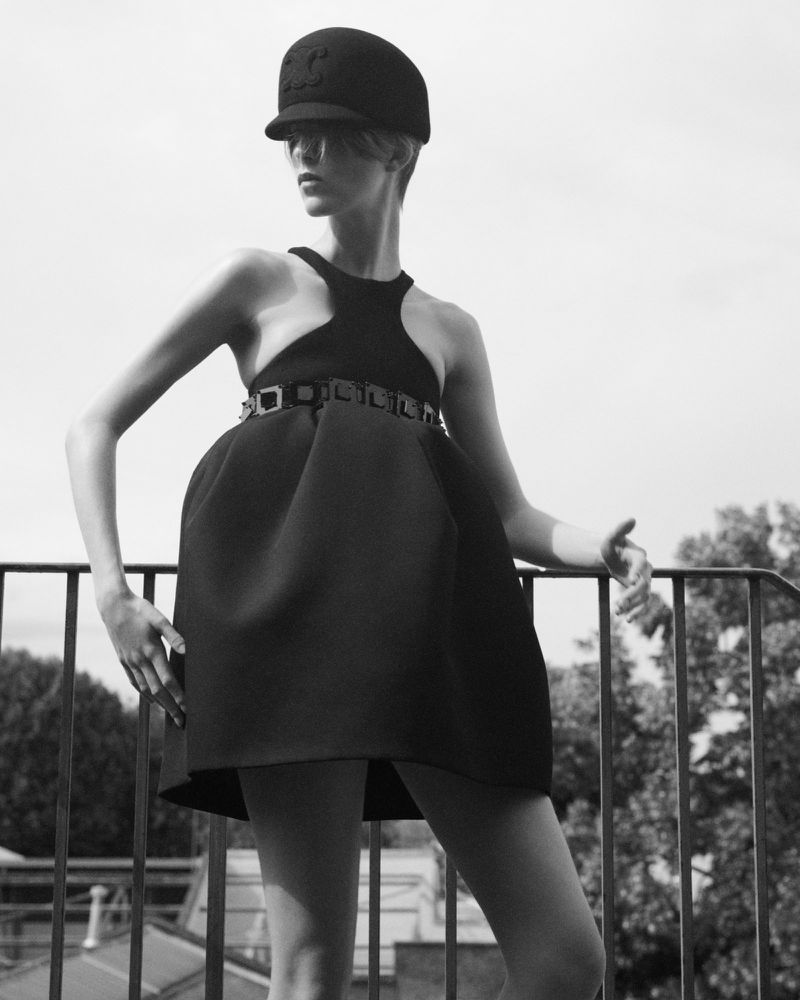 Black and white portrait of a young woman in a black dress and hat, posing outdoors against a background of sky and green trees.