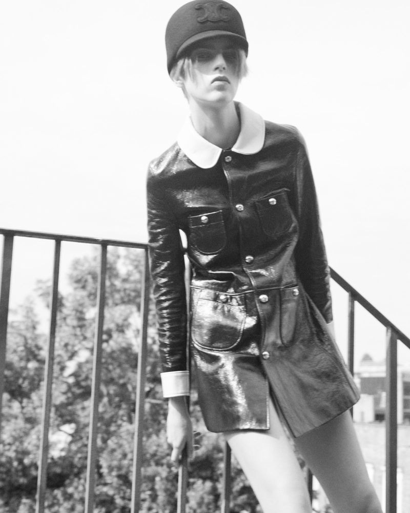 Black and white fashion shot of a young adult woman in a shiny black jacket and skirt, wearing a cap, standing on a balcony.
