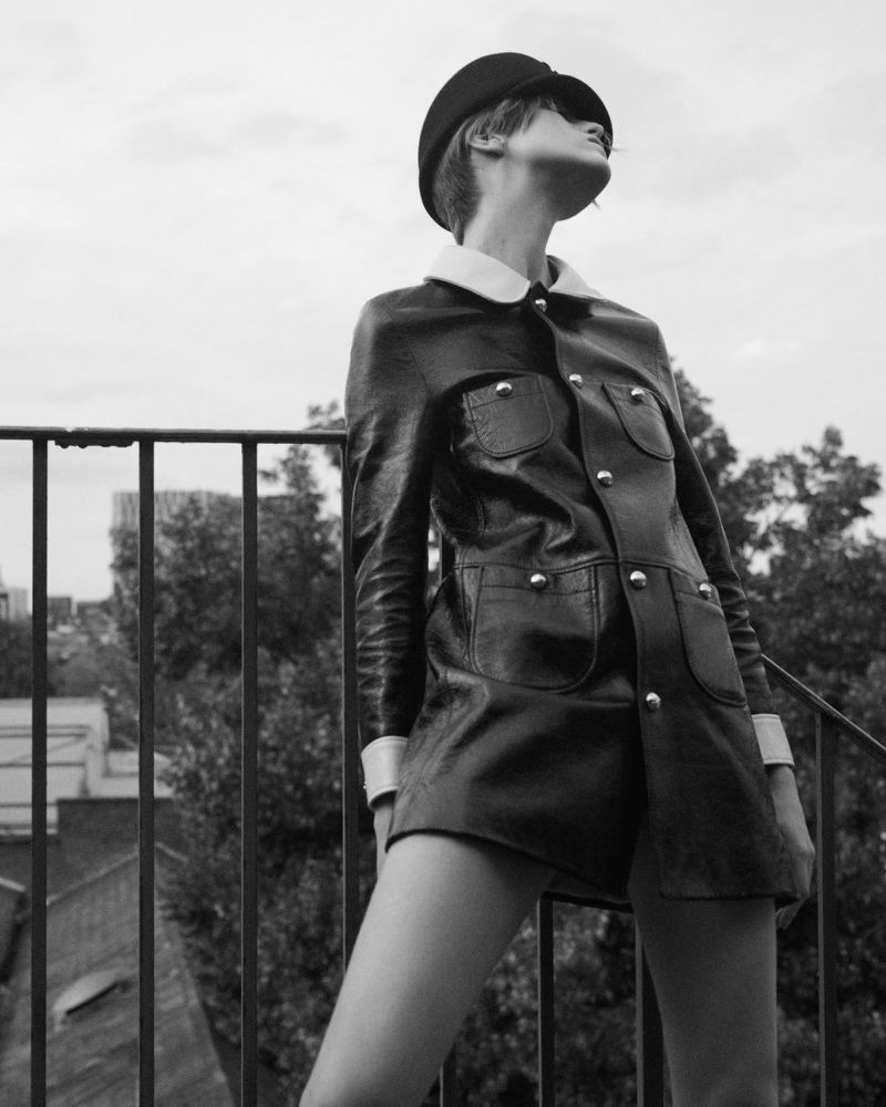 Black and white portrait of a woman in a vintage-style jacket and hat standing outdoors with trees and buildings in the distance.