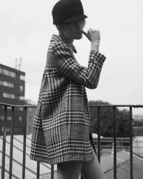 Model wearing a houndstooth coat and dark cap standing on a rooftop with a metal railing against an overcast sky