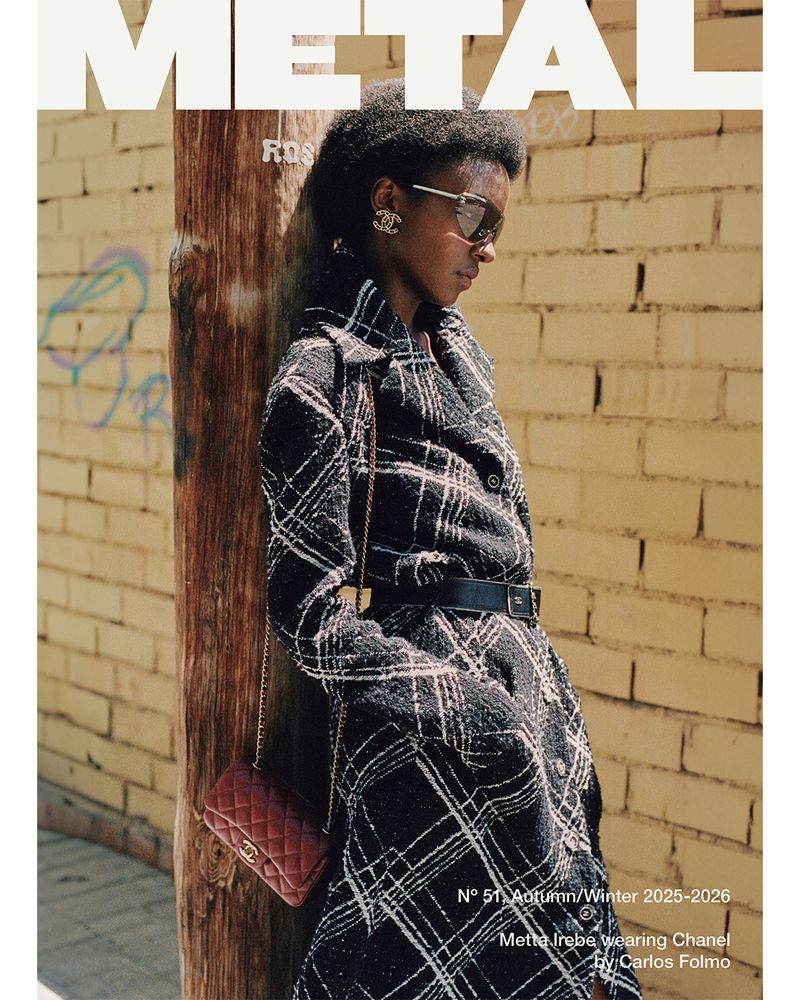 Editorial fashion shot of a Black woman wearing a plaid trench coat, sunglasses, and purse, posing against a weathered brick wall outdoors.