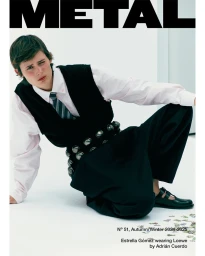 Model kneeling on a white studio floor wearing a black vest, white shirt, patterned tie, and wide trousers