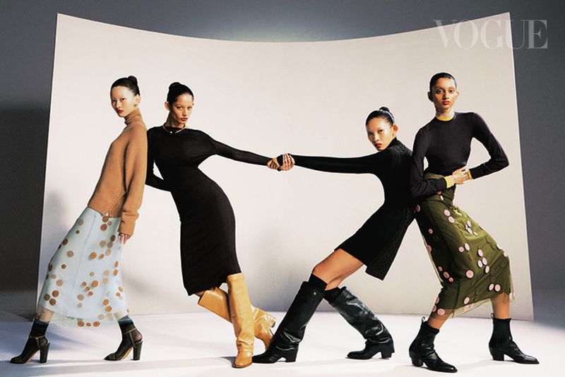 Four women in fashionable dresses and boots pose leaning against a curved white backdrop in a studio setting for a Vogue fashion shot.