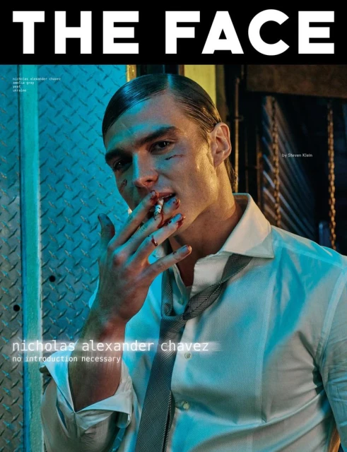 Magazine cover featuring a man with makeup and a cigarette, styled in a white shirt and tie, against a blue metal background.