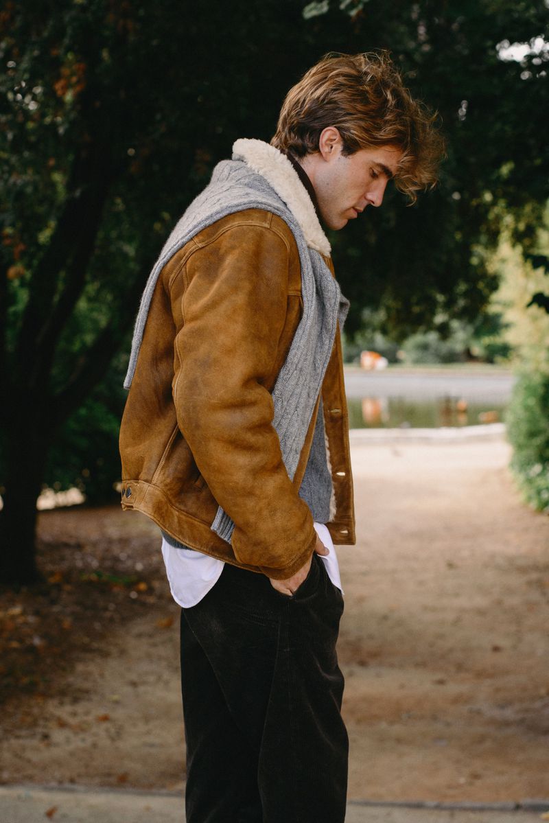 Adult male in a brown shearling jacket and grey scarf walks along a path with trees and a pond in the background, looking down.