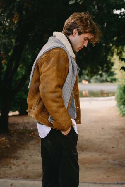 Adult male in a brown shearling jacket and grey scarf walks along a path with trees and a pond in the background, looking down.