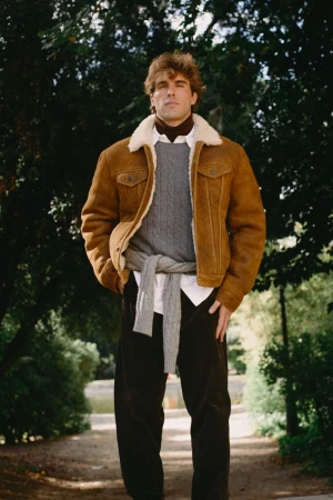 Man wearing a brown shearling collar jacket, grey cable-knit sweater, and dark trousers outdoors by a park path