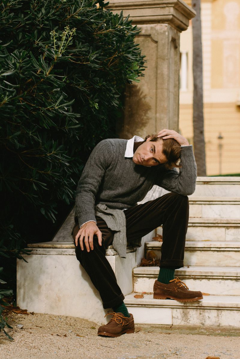 Adult male model in casual sweater, corduroy pants, and brown shoes poses seated on stone steps surrounded by trees and building exterior.