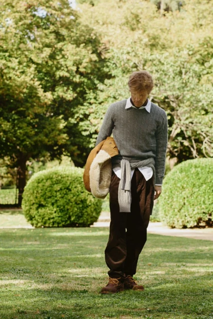 Full shot of a blond adult male standing on a grassy lawn wearing casual attire with a jacket over his shoulder in a park setting.