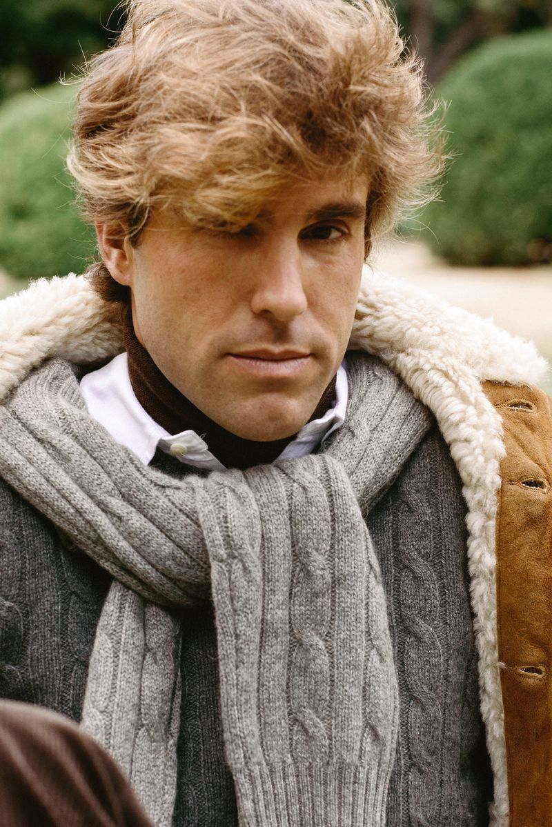 Man with wavy blond hair poses in a grey cable-knit scarf and brown coat with a shearling collar in an outdoor setting with green foliage.
