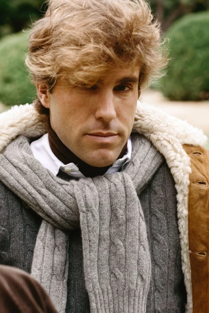 Man with wavy blond hair poses in a grey cable-knit scarf and brown coat with a shearling collar in an outdoor setting with green foliage.