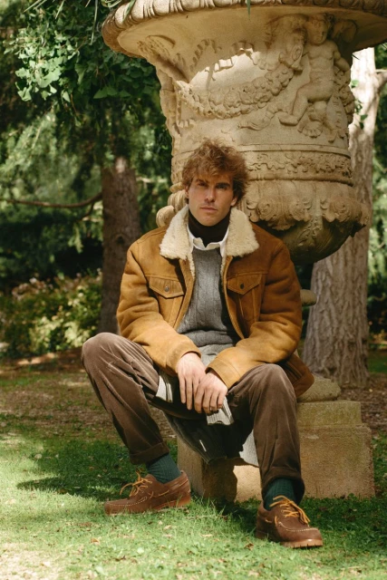 Adult male with curly hair sits near a decorative stone vase outdoors, wearing a brown sherpa jacket and corduroy pants.