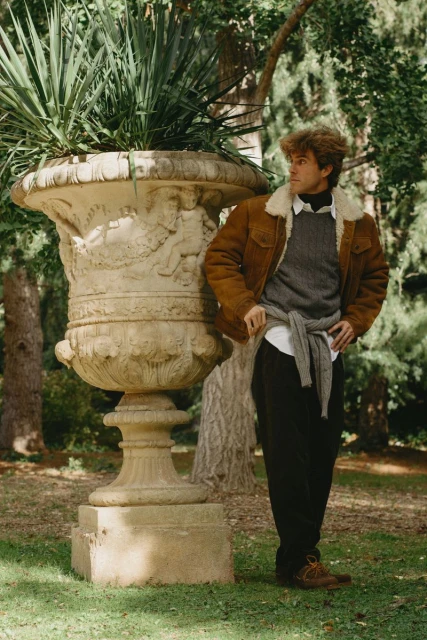 A young adult male poses next to a large stone planter outdoors in a park-like setting, wearing a brown jacket and dark pants.