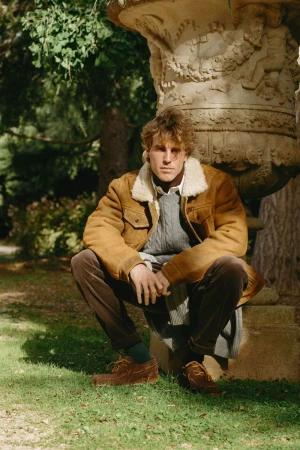 Man in a camel shearling jacket, grey cable knit sweater, and brown corduroy pants crouching by a stone urn in a garden.