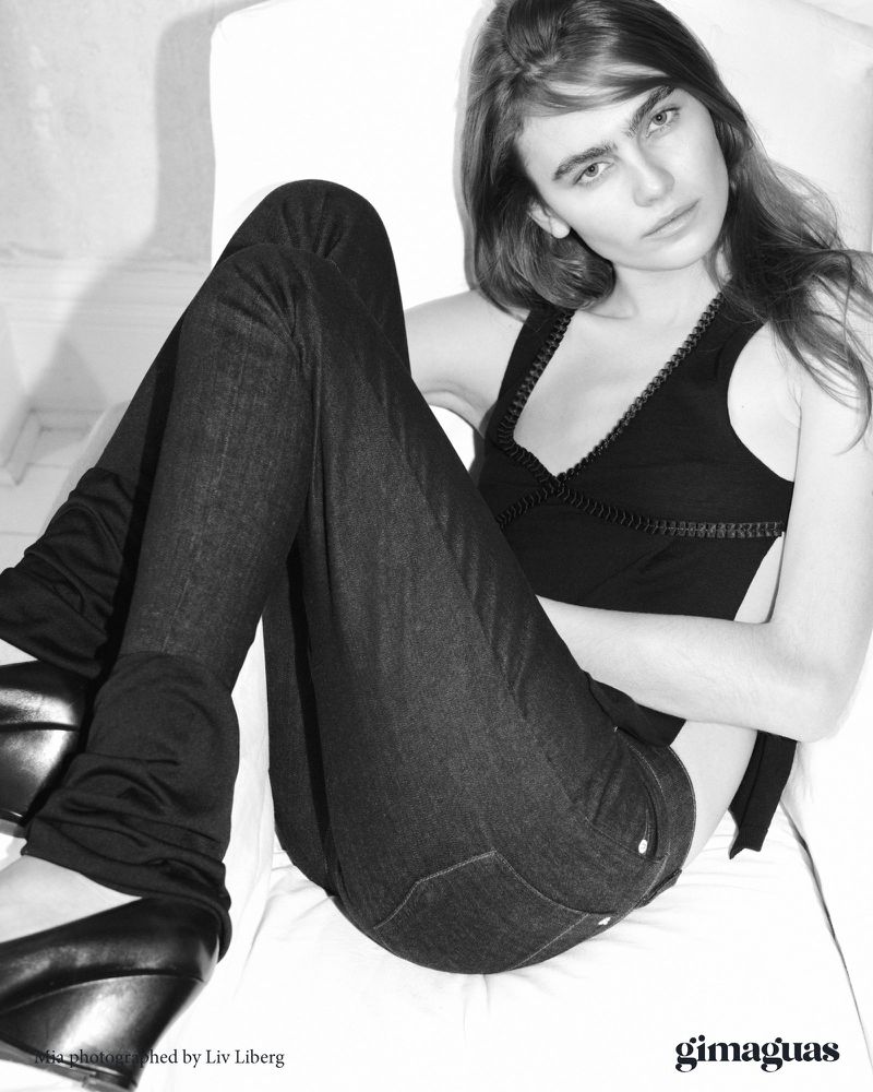 Monochrome shot of a young adult woman in a black top and jeans, sitting indoors on white fabric with dark wedge shoes.