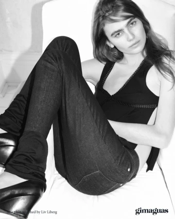 Monochrome shot of a young adult woman in a black top and jeans, sitting indoors on white fabric with dark wedge shoes.