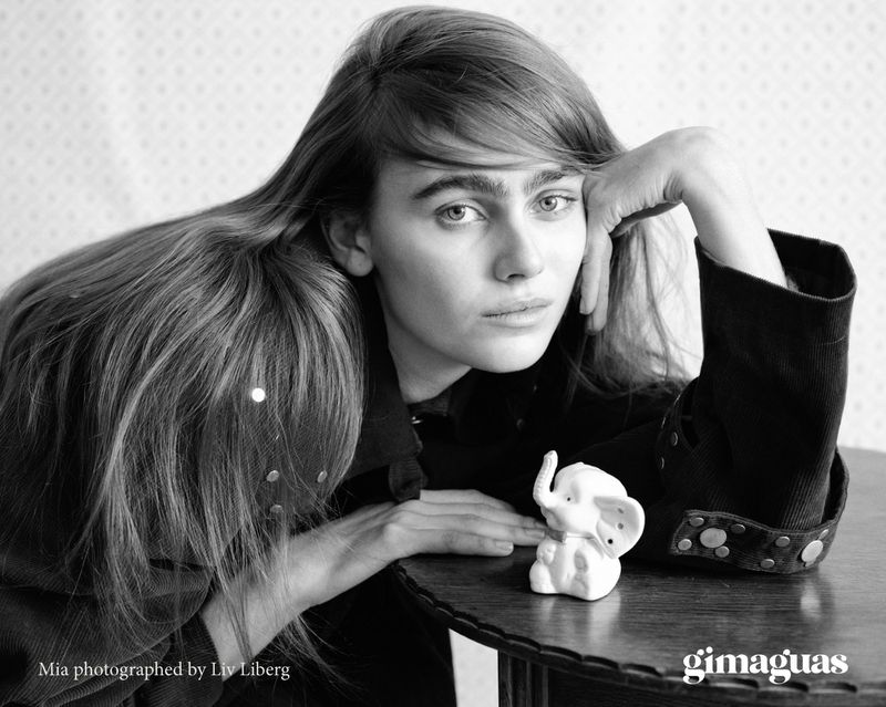Black and white portrait of woman with long hair resting on table next to small elephant figurine in front of patterned wallpaper.