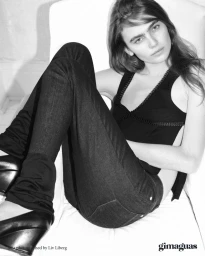 Mia Armstrong reclining on a white chair wearing dark fitted jeans, a black sleeveless top, and black wedge heels.