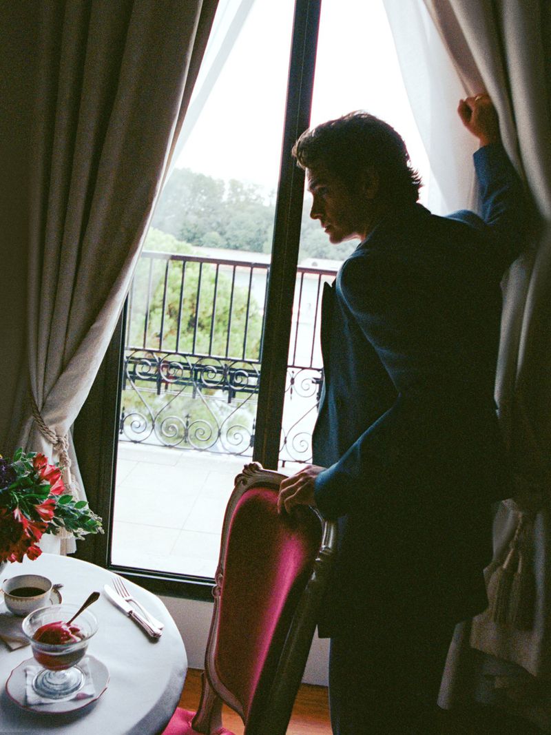 A man in a suit stands near a window with curtains, a table with a dessert and flowers are also visible in an indoor setting.