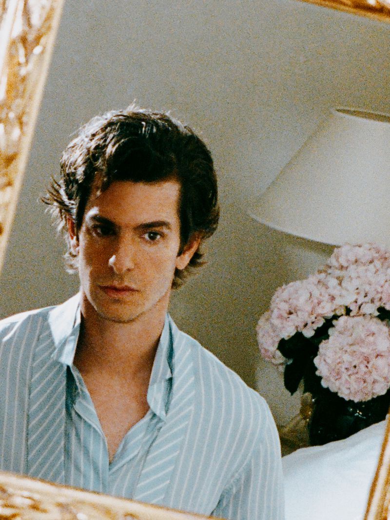 Andrew Garfield wearing a light blue striped shirt reflected in an ornate gold mirror beside pink flowers.
