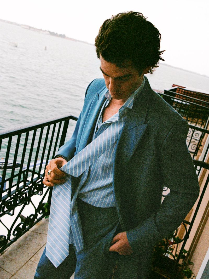 Portrait of a handsome adult man in a stylish blue suit standing on a balcony overlooking the ocean with muted sunlight. 