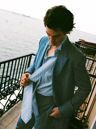 Portrait of a handsome adult man in a stylish blue suit standing on a balcony overlooking the ocean with muted sunlight. 