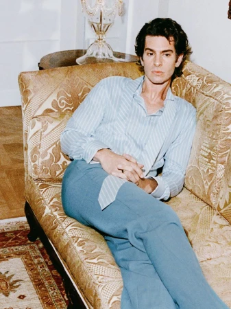 Andrew Garfield poses on a vintage couch in a photo studio. He is wearing a light blue shirt and blue pants.