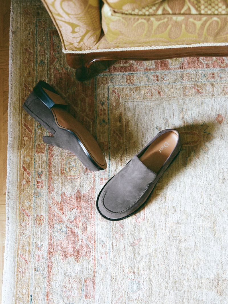 Two grey suede slip-on shoes displayed on an ornate rug, with a hint of a patterned armchair visible in the background.